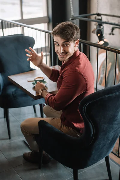 Joven Sonriente Sentado Mesa Madera Con Capuchino Mano Agitada Balcón — Foto de Stock