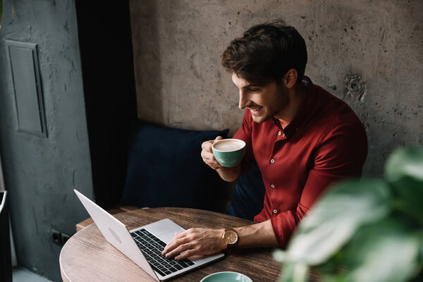smiling young freelancer working on laptop and drinking coffee in coffee shop