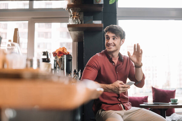 smiling handsome man sitting near bar counter in wireless earphones with smartphone and waving hand