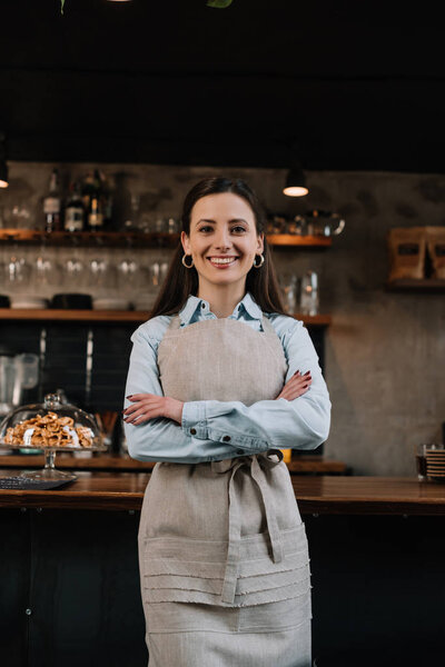 smiling barista in apron standing with crossed arms near bar counter