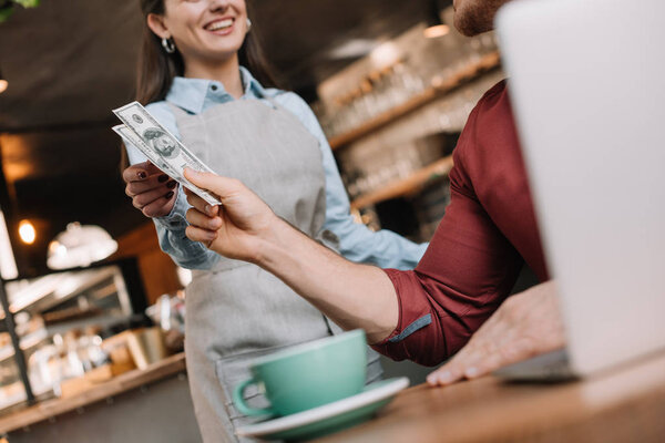 cropped view of freelancer with laptop giving cash to smiling waitress in coffee shop
