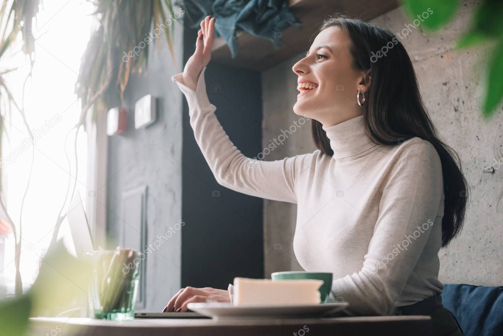 selective focus of green plants and happy freelancer using laptop and waving hand in coffee shop