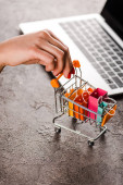 cropped view of woman holding toy shopping cart with small shopping bags near laptop, e-commerce concept