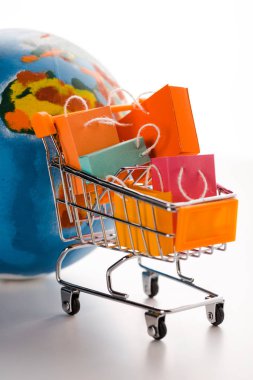 Beyaz, e-ticaret kavramı üzerine dünya yakınında küçük renkli alışveriş çantaları olan oyuncak alışveriş arabası.
