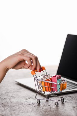 Beyaz, e-ticaret konseptine izole edilmiş, dizüstü bilgisayarın yanında küçük alışveriş çantaları taşıyan oyuncak alışveriş arabası tutan bir kadın görüntüsü.