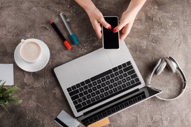 Laptop, kahve, bitki, kulaklık, kredi kartı ve e-ticaret konsepti yanında akıllı telefonu tutan kadının üst görüntüsü.