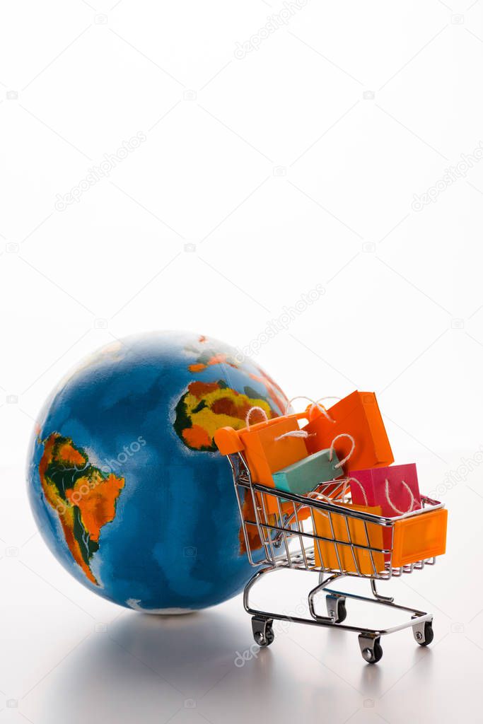 selective focus of toy shopping cart with shopping bags near globe on white, e-commerce concept