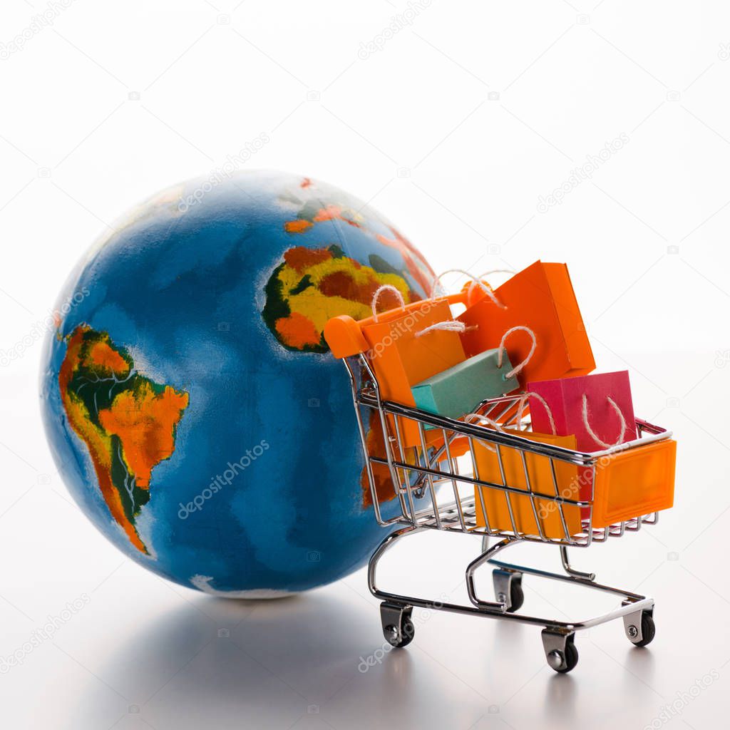 toy shopping cart with shopping bags near globe on white, e-commerce concept
