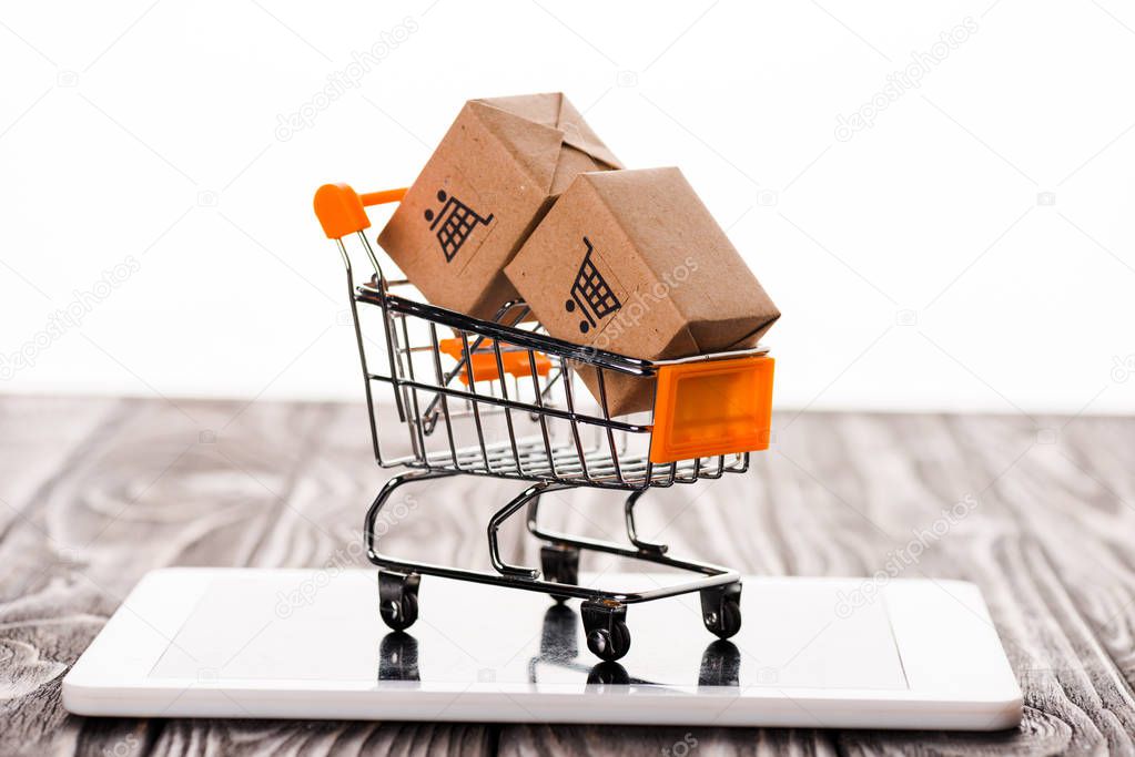 toy shopping cart with small carton boxes on digital tablet isolated on white, e-commerce concept