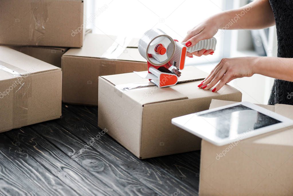 cropped view of woman holding adhesive tape near carton box and digital tablet in office, e-commerce concept