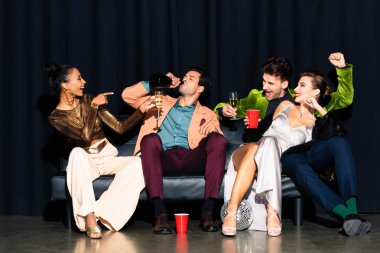happy african american girl pointing with finger at man drinking champagne from bottle near friends on dark blue