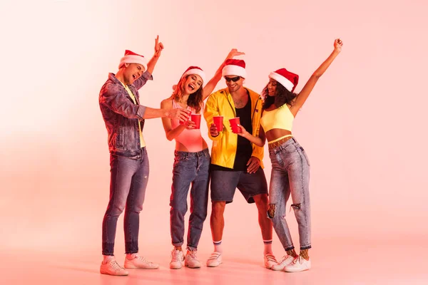 Aufgeregte Männer Und Multikulturelle Mädchen Weihnachtsmützen Mit Plastikbechern Auf Rosa — Stockfoto