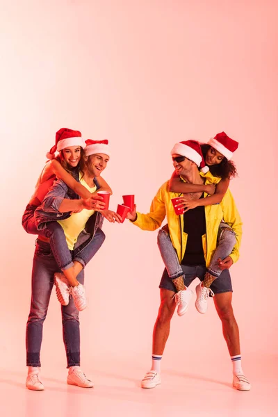 Happy Men Piggybacking Multicultural Girls Santa Hats Clinking Plastic Cups — Stock Photo, Image