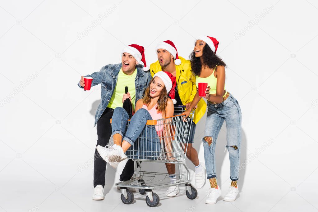happy girl sitting in shopping cart with bottle near multicultural friends with plastic cups on white 