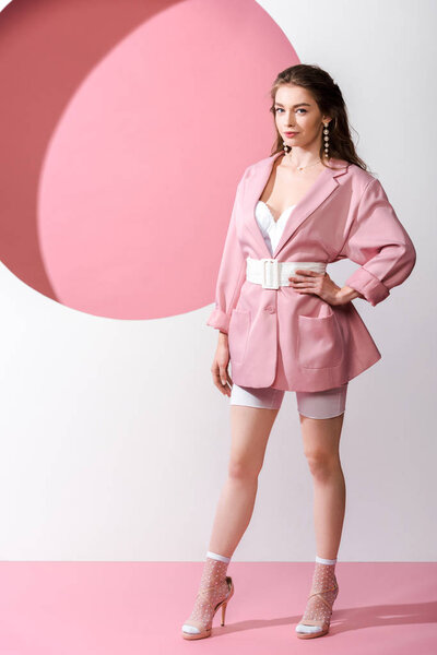 stylish woman in blazer with belt standing with hand on hip on white and pink 