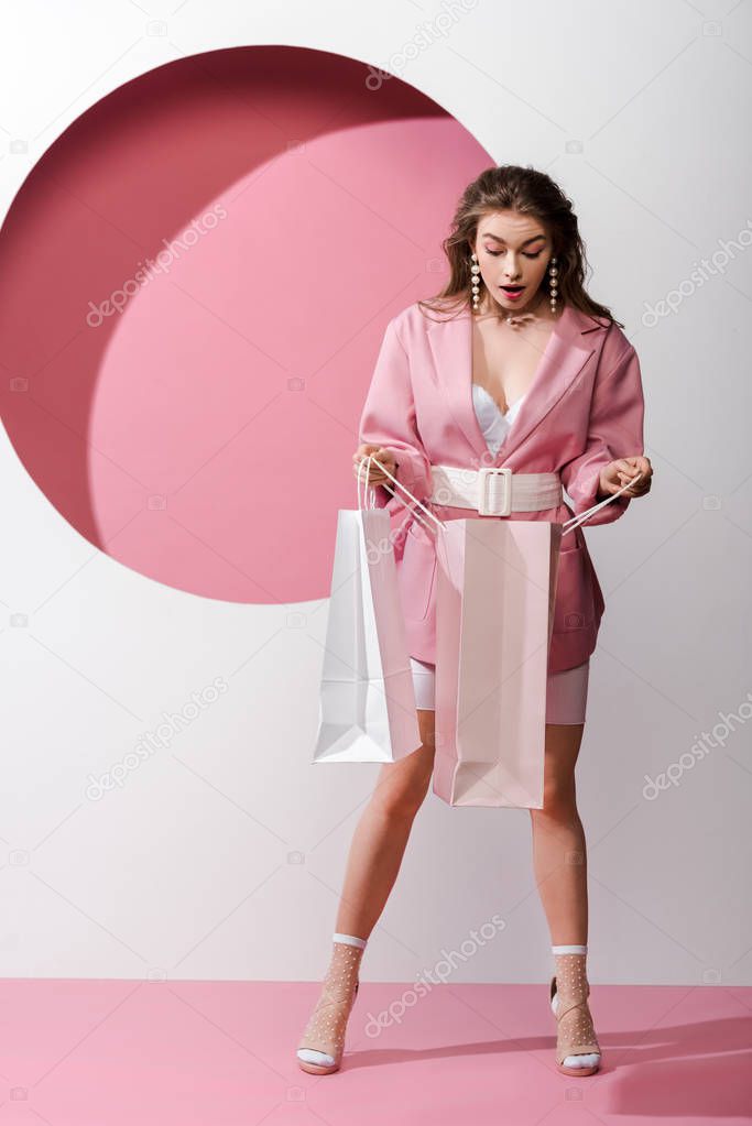 surprised and stylish girl looking at shopping bag on white and pink 