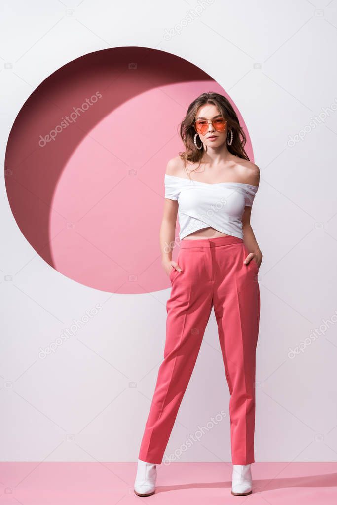 attractive girl in sunglasses standing with hands in pockets on pink and white 