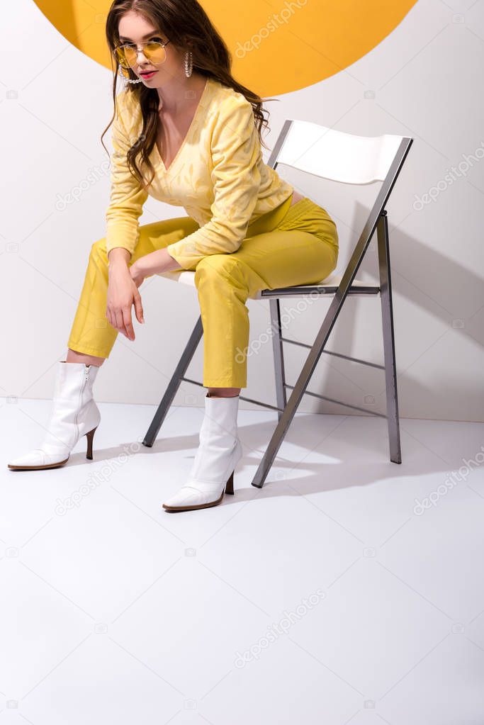 attractive and stylish woman in sunglasses sitting on chair on white 