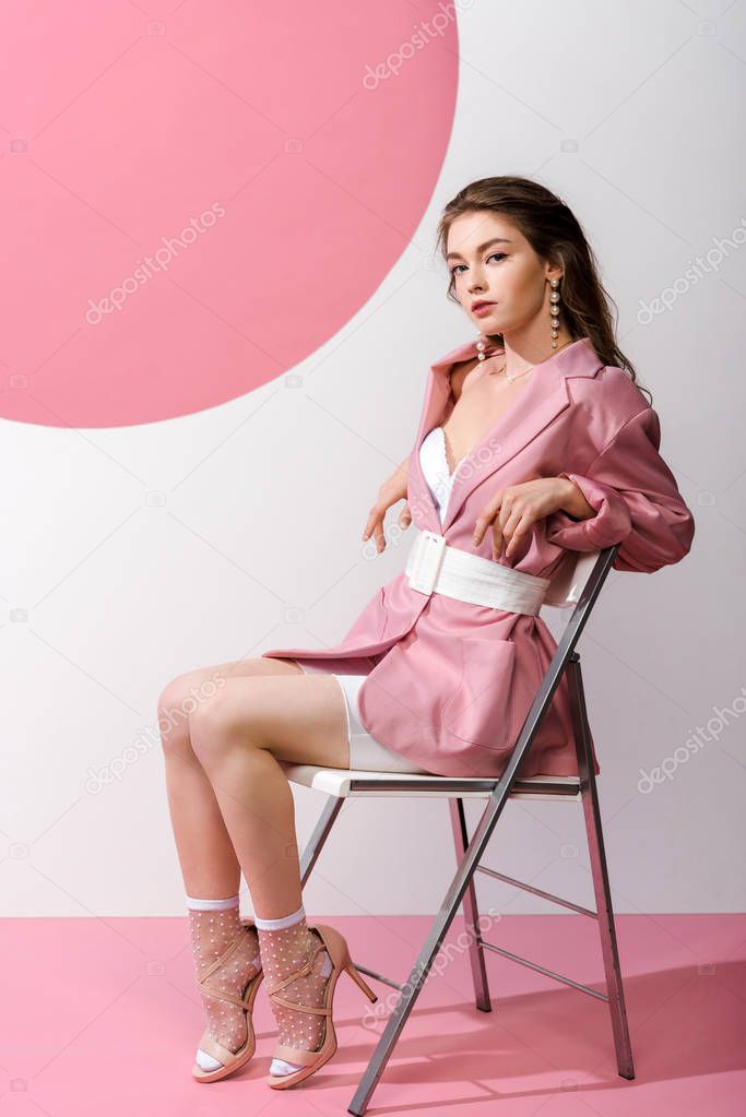 stylish young woman in blazer with belt sitting on chair on white and pink 