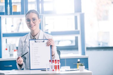 attractive and happy immunologist holding clipboard with checklist and pen  clipart