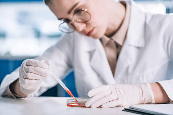 selective focus of attractive immunologist in eye glasses holding pipette with red liquid in laboratory 