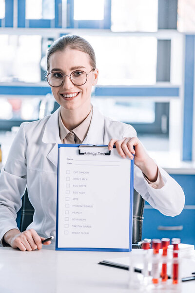 attractive and cheerful immunologist holding clipboard with checklist and pen near test tubes