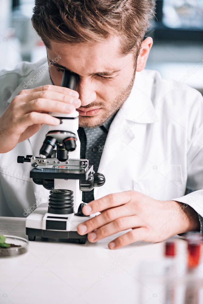 selective focus of handsome biochemist looking through microscope in laboratory 