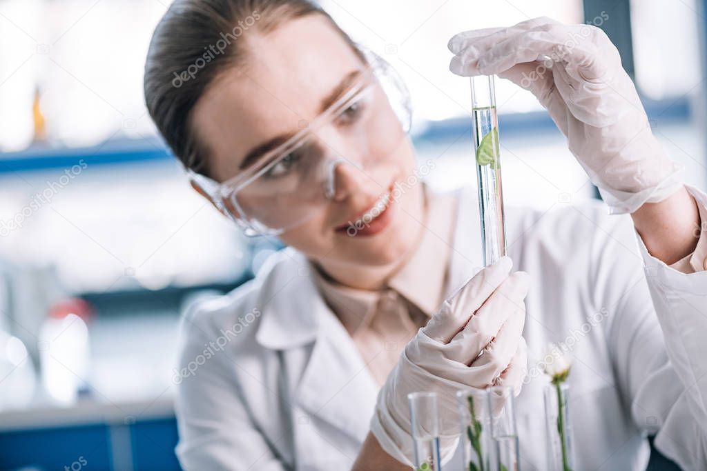 selective focus of cheerful biochemist in goggles holding test tube with small green plant