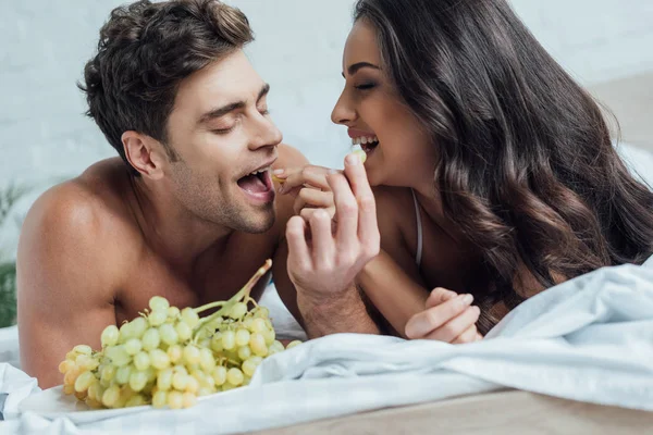幸せな若いカップルはベッドの上に横になりながらブドウを食べます — ストック写真
