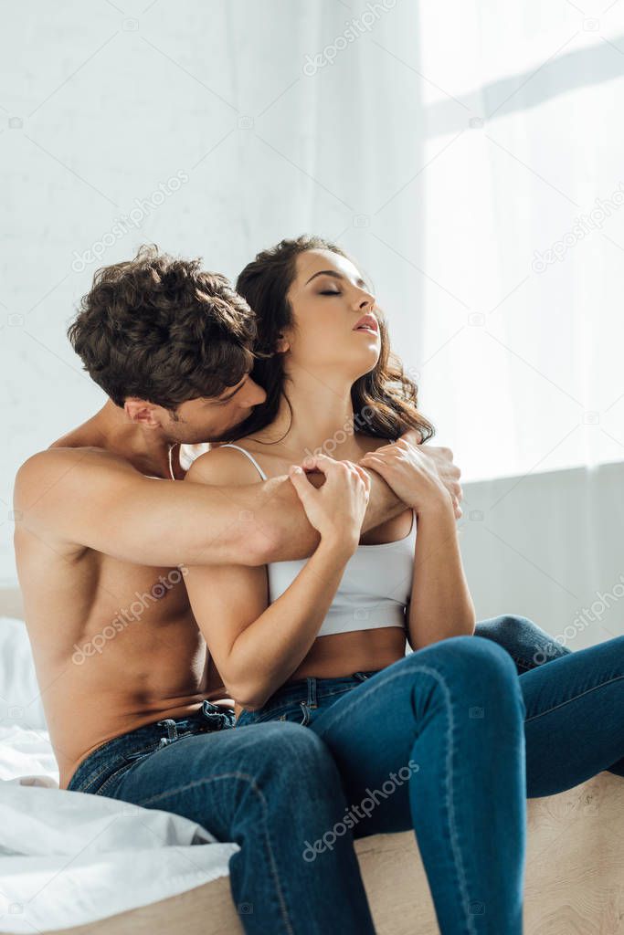shirtless man kissing and hugging sexy girlfriend while sitting on bed 