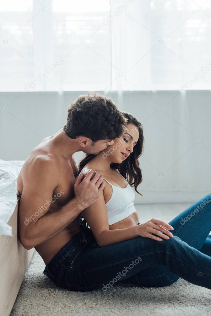 shirtless man kissing and hugging girlfriend while sitting on floor