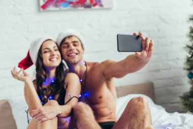 İç çamaşırlı ve Noel Baba şapkalı neşeli bir çift yatakta akıllı telefondan selfie çekiyor.