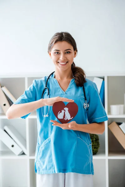 Attractive Positive Veterinarian Smiling Camera While Presenting Paper Cut Heart — Stock Photo, Image