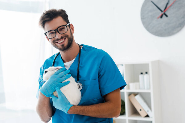 handsome, positive veterinarian smiling at camera while holding cute black and white rabbit on hands