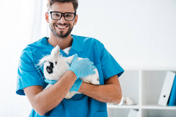 young, positive veterinarian smiling at camera while holding cute black and white rabbit on hands