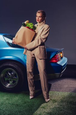 attractive and stylish woman in suit standing near retro car and holding paper bag with food  clipart
