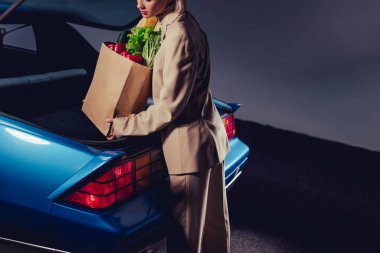 cropped view of stylish woman in suit packing paper bag with food in trunk  clipart