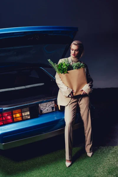 穿着西服的迷人而时髦的女人站在复古汽车旁边 拿着装有食物的纸袋 — 图库照片