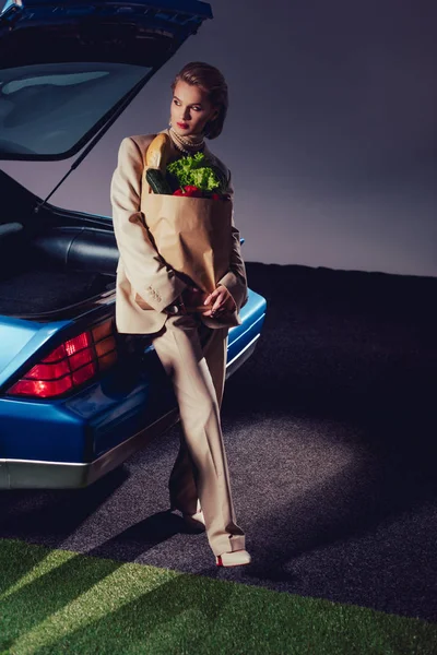 レトロな車の近くに立つスーツ姿の魅力的でスタイリッシュな女性食品と紙袋を保持 — ストック写真