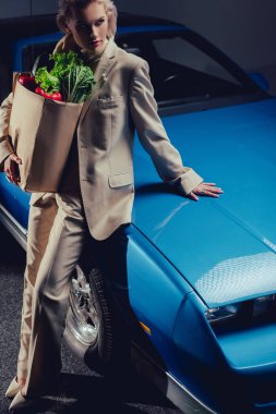 high angle view of attractive and stylish woman in suit standing near retro car and holding paper bag with food clipart