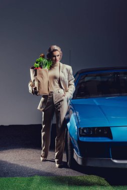 attractive and stylish woman in suit standing near retro car and holding paper bag with food  clipart
