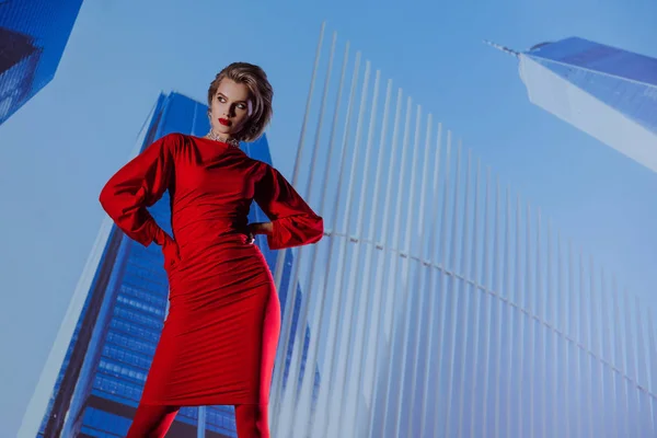 魅力的でスタイリッシュな女性の低角度ビュー都市の背景に赤いドレスで腰に手 — ストック写真