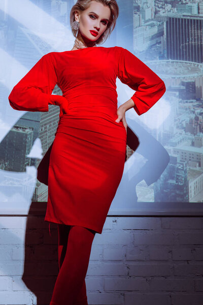 attractive and stylish woman in red dress with hands on hips on city background 