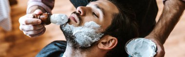 panoramic shot of barber applying shaving cream on face of bearded man  clipart