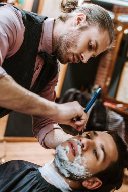 selective focus of handsome barber shaving bearded man with shaving cream on face  clipart