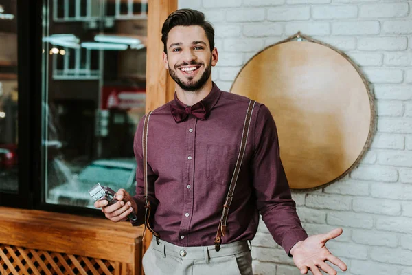 happy bearded barber holding trimmer and gesturing in barbershop