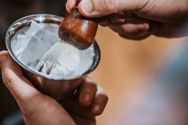 cropped view of barber holding shaving brush near bowl with white shaving cream 