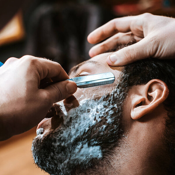 close up of barber shaving bearded man with shaving cream on face 