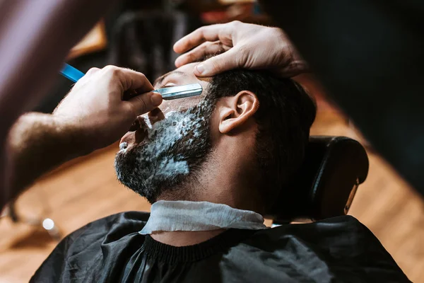 selective focus of barber shaving man with shaving foam on face