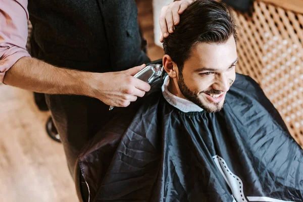 barber holding trimmer while styling hair of happy man in barbershop